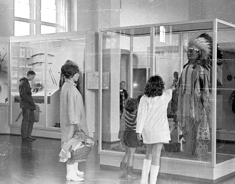 View into the permanent exhibition in the North America department – © MARKK Showcases, dressed mannequin, exhibition visitors, rubber boots, b/w photograph